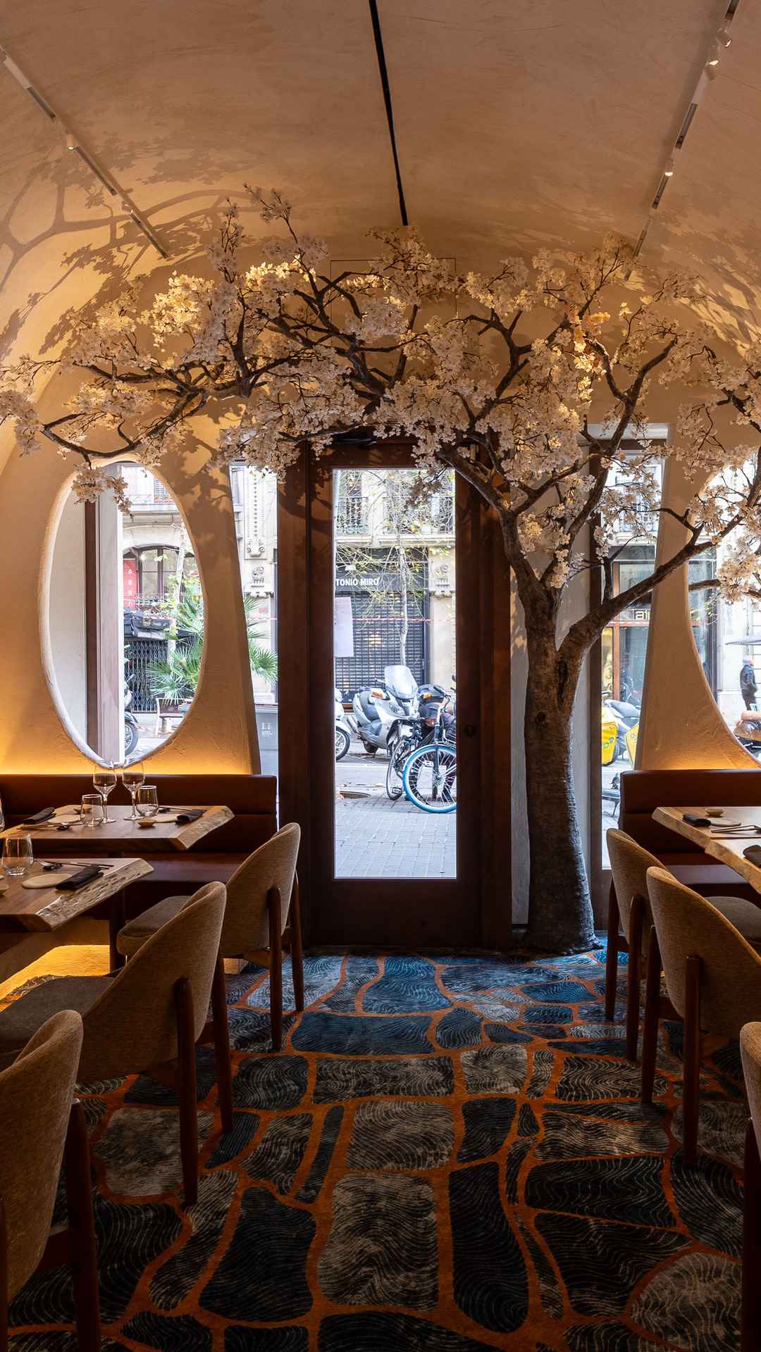 La entrada de Arko: un cerezo en flor sobre la puerta de cristal, ventanas ovaladas y mesas de nogal